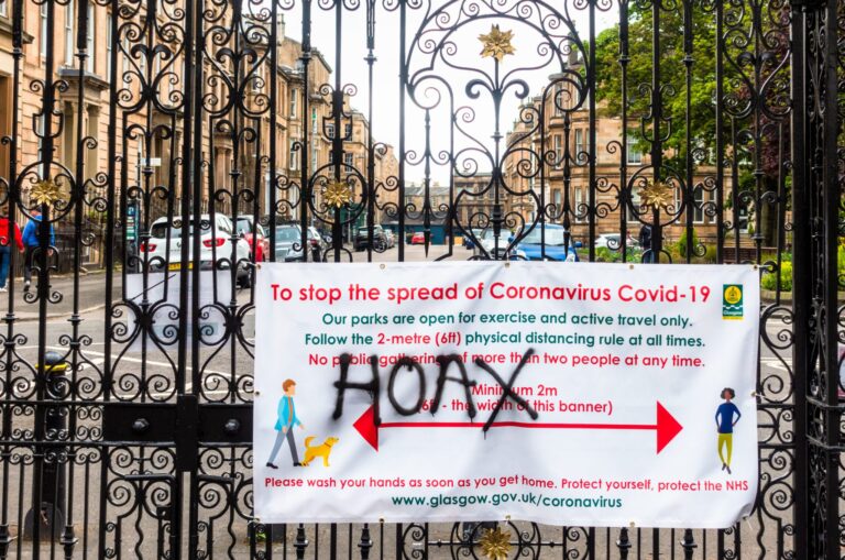 The word 'hoax' spray painted onto a banner sign communicating information about reducing the spread of covid-19 pandemic